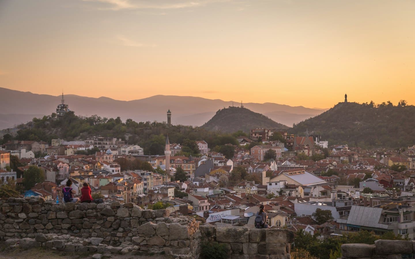 Dos personas sentadas en un muro de piedra con vistas a una ciudad histórica.